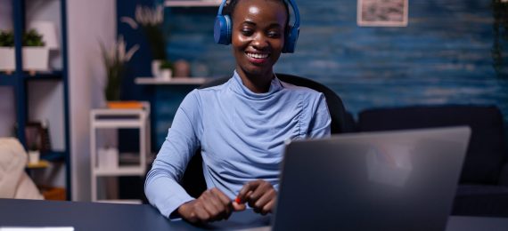 Black woman in good mood wearing headset listening music working on deadline from home office. sitting at desk. African freelancer creating new project working late.