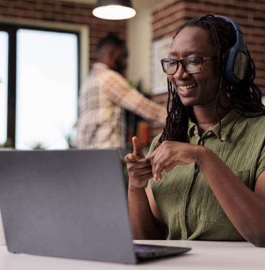 Smiling woman with wireless headphones watching entertainment show on laptop while sitting at desk. African american freelancer looking at funny social media content on portable computer.